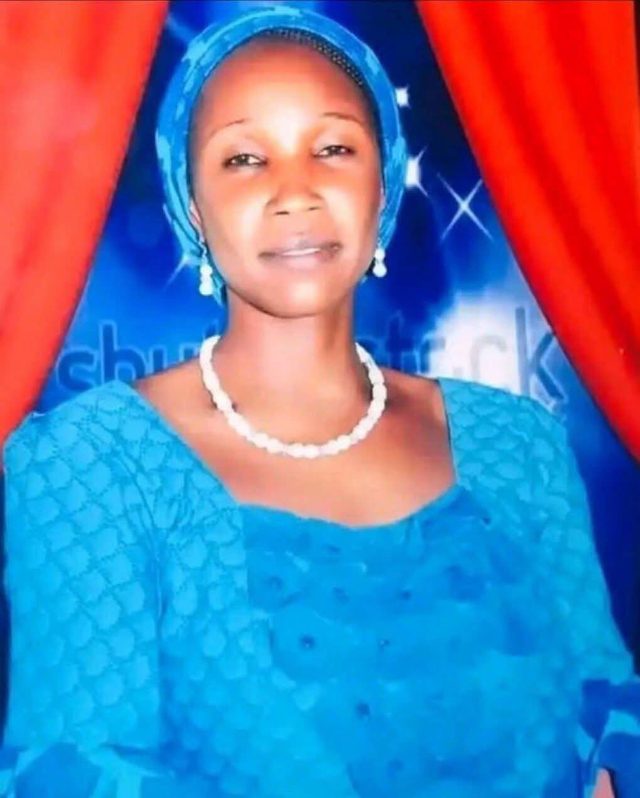 Portrait of Liyatu Samuel Dawa in a blue headscarf and blue dress with a white bead necklace, in front of red curtains