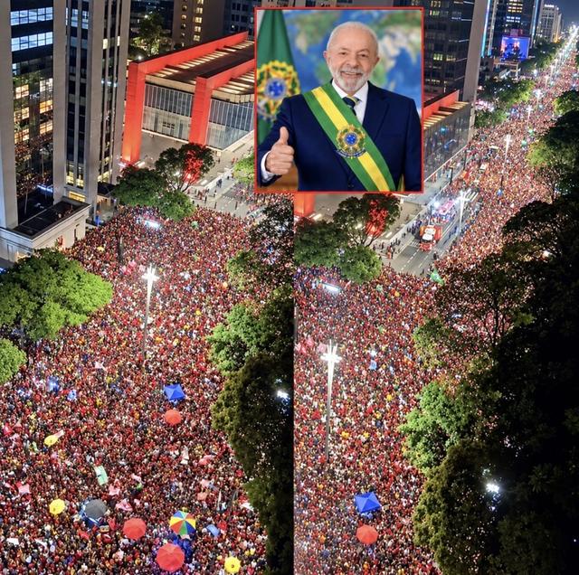 Aerial view of a huge crowd in red filling a wide city avenue at night, with an inset photo of Lula in the presidential sash giving a thumbs-up