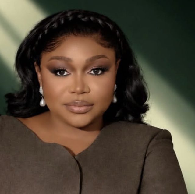Ruth Kadiri in a brown jacket and pearl earrings against a dark green background