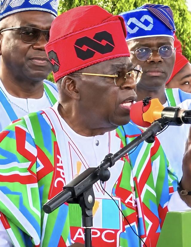 President Bola Tinubu, in a red cap and APC-branded attire, speaks into a microphone as Governor Babajide Sanwo-Olu and another official look on