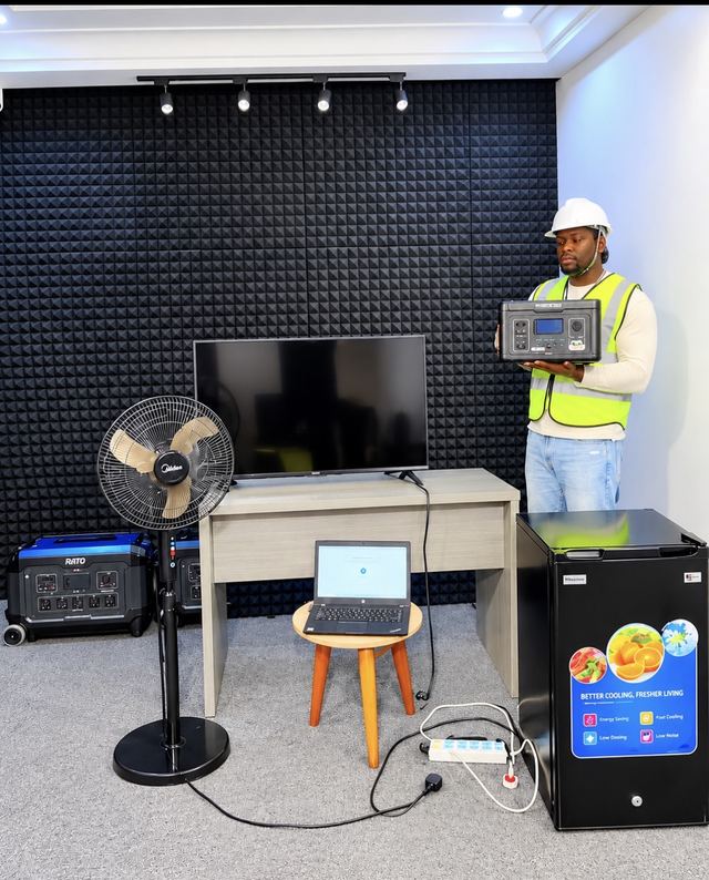 VeryDarkMan in a hard hat and hi-vis vest holds a Ratel power station beside a TV, standing fan, laptop and fridge set up for a test